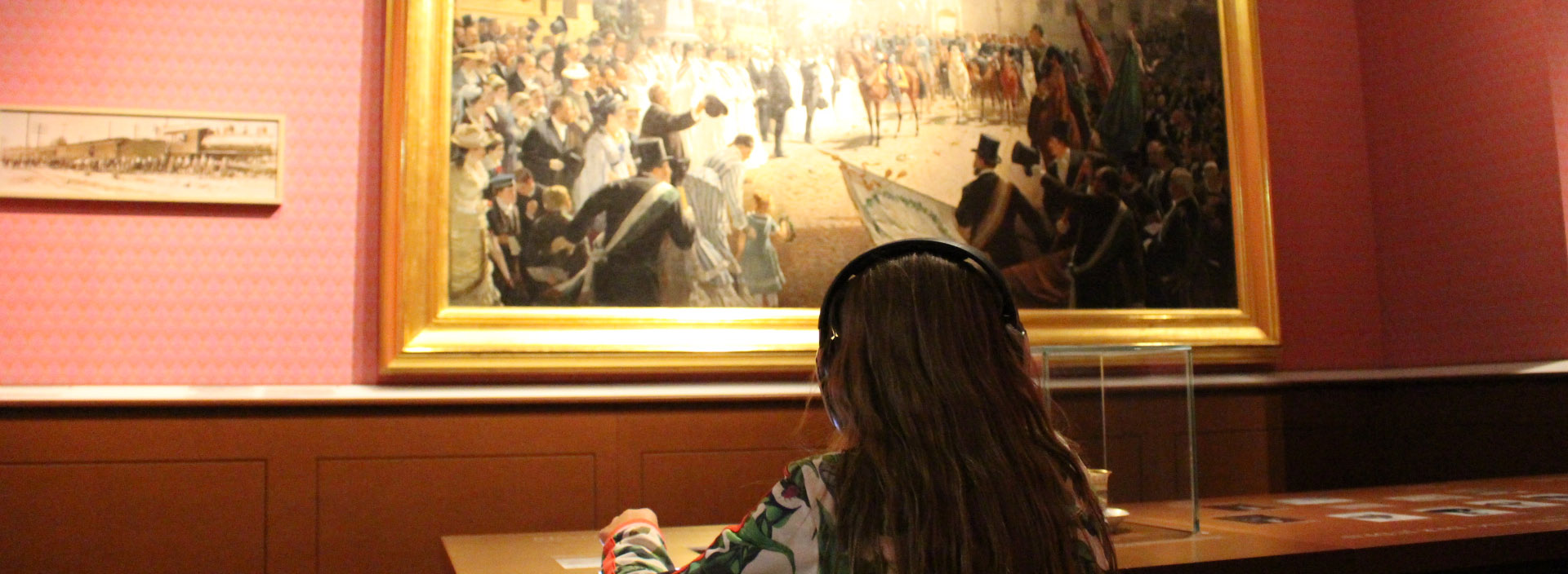 Ein Mädchen sitzt mit Kopfhörern am Tisch und betrachtet dabei ein historisches Gemälde.